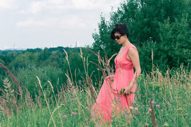 A middle-aged woman in a pink dress is walking in the park. The city is on the horizon. The concept is a rest in city parks.