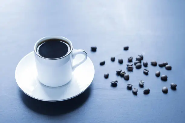 A white cup of fragrant hot coffee on the morning table. Simple colors and grains of roasted coffee.