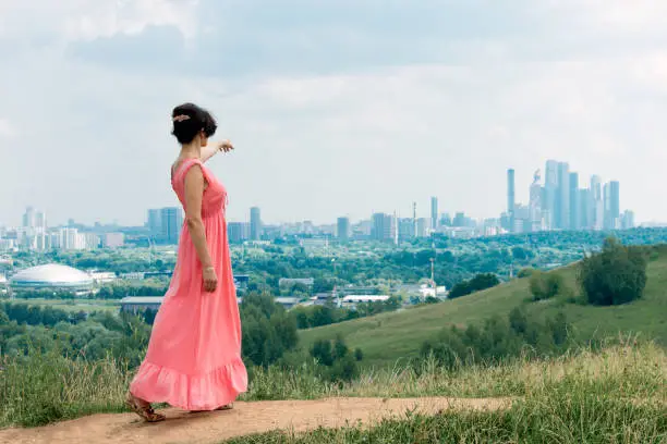 Concept - life in a big city and nature. A middle-aged woman walks along a path from a hill and points to a city. On the horizon, Moscow, Russia.