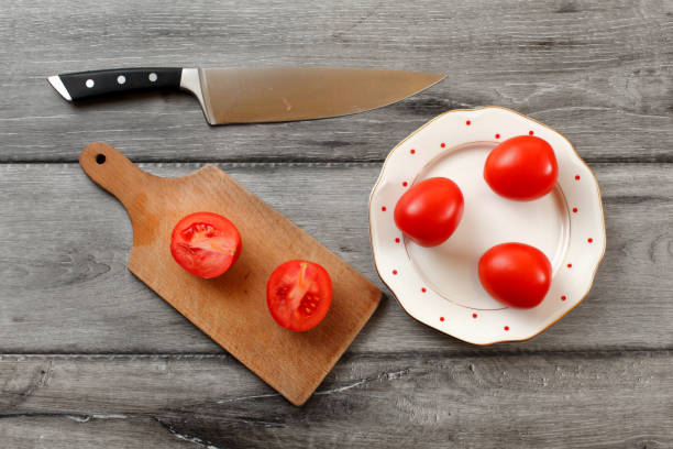 istock Table top view on three tomatoes on white porcelain plate with red dots, cutting board with tomato cut to half next to it, chef knife also laying on the gray wood desk. 1007144402