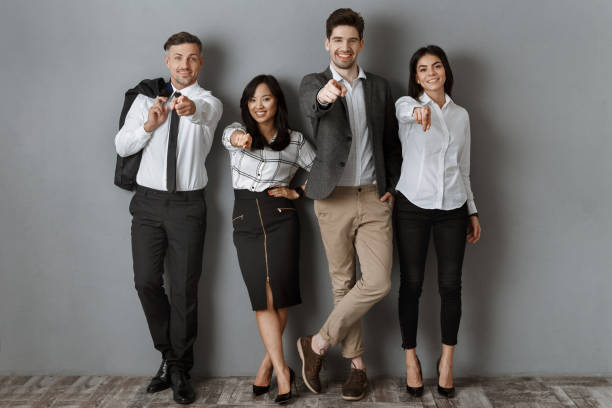 istock multicultural business people in formal wear pointing at camera while standing at grey wall 1007147466
