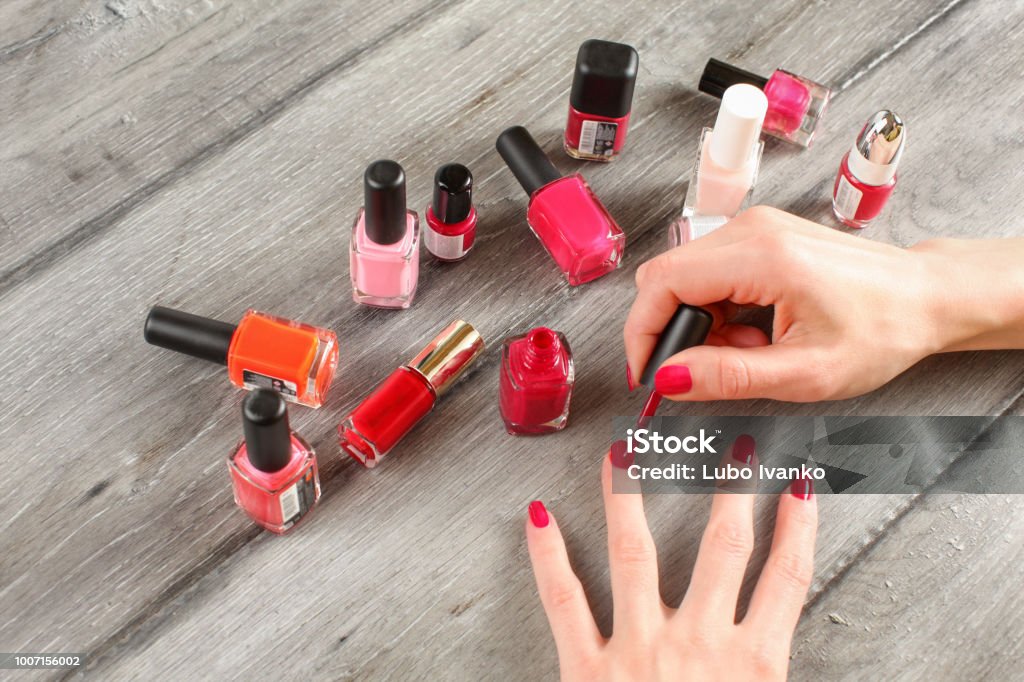 Top View On Young Woman Hands Applying Second Red Coat Of Nail Varnish With Glass Bottles With Nail Polish In The Background On Gray Wood Desk Stock Photo - Download Image Now - Top View On Young Woman Hands Applying Second Red Coat Of Nail Varnish With Glass Bottles With Nail Polish In The Background On Gray Wood Desk Stock Photo - Download Image Now -