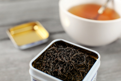 Black loose tea in metal tin, with blurred cup of hot amber drink in background.