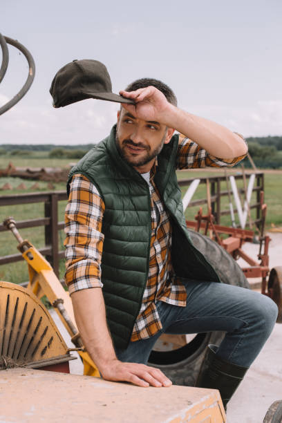 istock handsome male farmer holding cap and looking away while working at ranch 1007188996