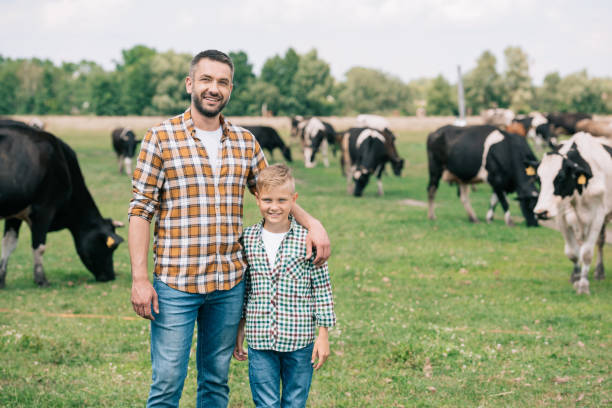 padre e figlio sorridente alla macchina fotografica mentre si trova vicino al bestiame al pascolo in fattoria - farmer farm family son foto e immagini stock