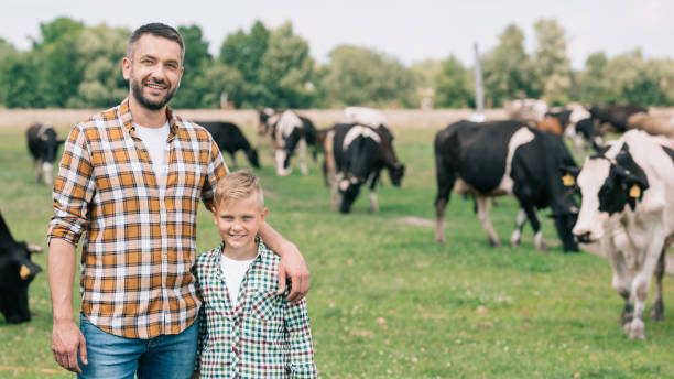 happy father and son smiling at camera while standing near grazing cattle at farm - boyhood imagens e fotografias de stock