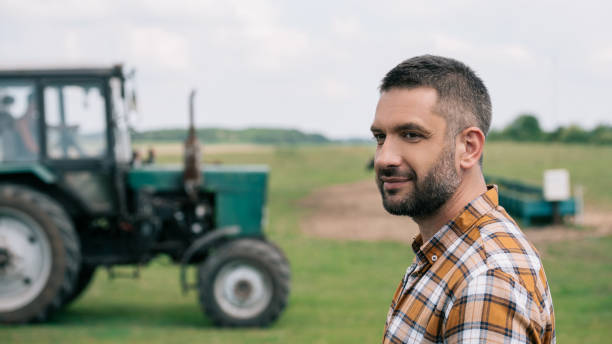 istock side view of handsome middle aged farmer smiling while standing near tractor in field 1007189106