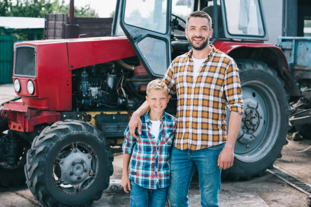 happy father and son in checkered shirts smiling at camera while standing together neat tractor at farm - boyhood imagens e fotografias de stock