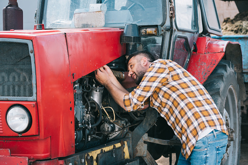 handsome middle aged farmer in checkered shirt repairing tractor