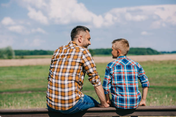 back view of father and son sitting on fence and smiling each other at farm - boyhood imagens e fotografias de stock