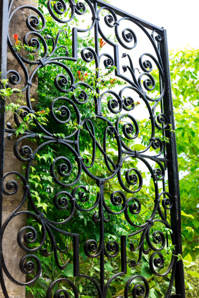 Vertical color image depicting a black wrought iron garden gate with lush green foliage beyond the gate. The gate is open, providing an invitation into the garden. Room for copy space.