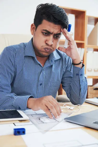 Tired Indian office worker sitting at desk and breathing out while touching head