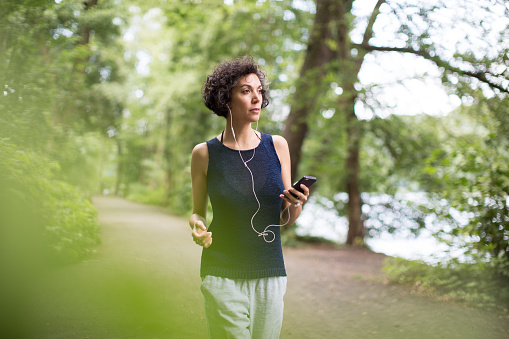 Woman listening to music on in-ear headphones using mobile phone. Confident mature female is walking on road. She is wearing casuals in forest.