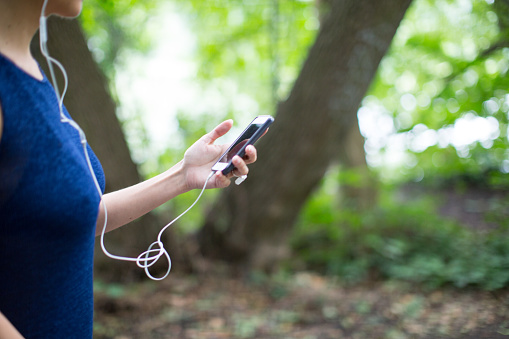 Cropped image of mature woman using smart phone and in-ear headphones in forest. Midsection side view of female is hiking in woodland. She is enjoying during vacation.