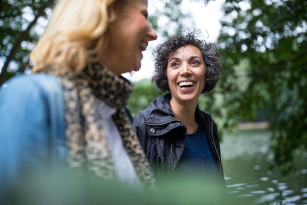 Happy mature woman looking at friend in forest Happy mature woman looking at friend in forest. Female friends are sitting at lakeshore. They are communicating in woodland. selective focus stock pictures, royalty-free photos & images