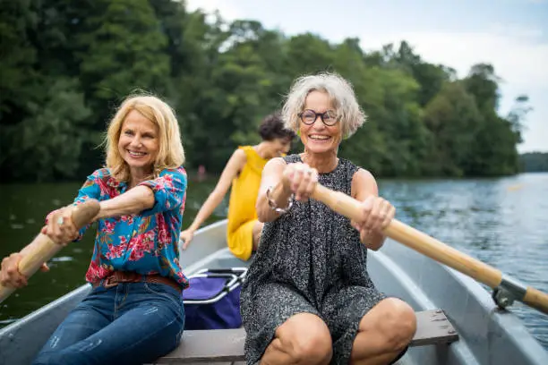 Smiling female friends rowing boat in lake. Mature women are enjoying boating in forest. They are on vacations.