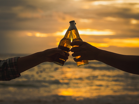 Couple hands holding beer bottles and clanging on the sunset beach. Party, Holiday, Summer, Friendship Concept.