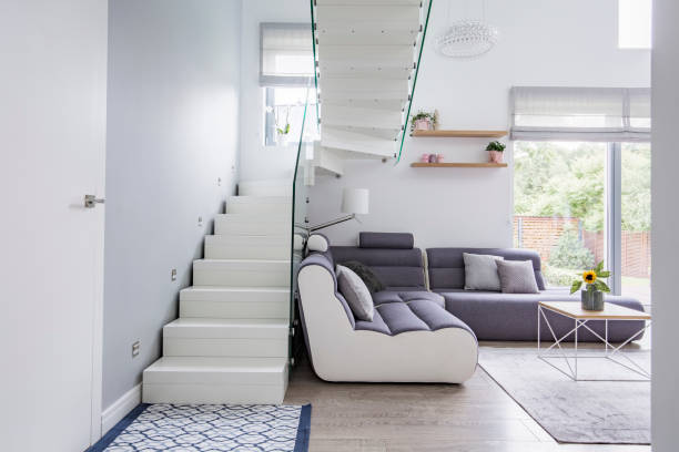 istock Large, modern sofa in a white living room interior with natural light and stylish staircase with glass barricade 1007254270