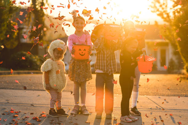 Multiethnic group of kids trick or treating Five children of varying ages, ethnicities, and costumes go trick or treating. They include a cowboy, a duck, a fitness instructor, a gymnast, and a black cat. They are holding candy buckets in a residential neighborhood and they're throwing leaves in the air. trick or treat photos stock pictures, royalty-free photos & images