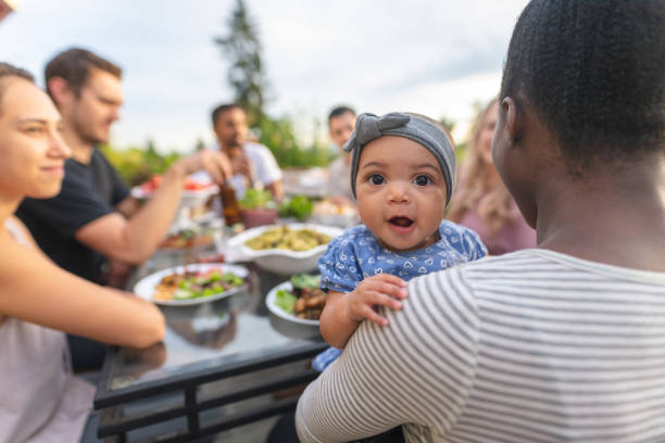 A group of young adult friends dining al fresco on a patio A multiethnic group of young friends enjoy good food and conversation together on a terrace outside on a summer evening. The focus is on an adorable young girl who's smiling at the camera while mom holds her. picnic photos stock pictures, royalty-free photos & images