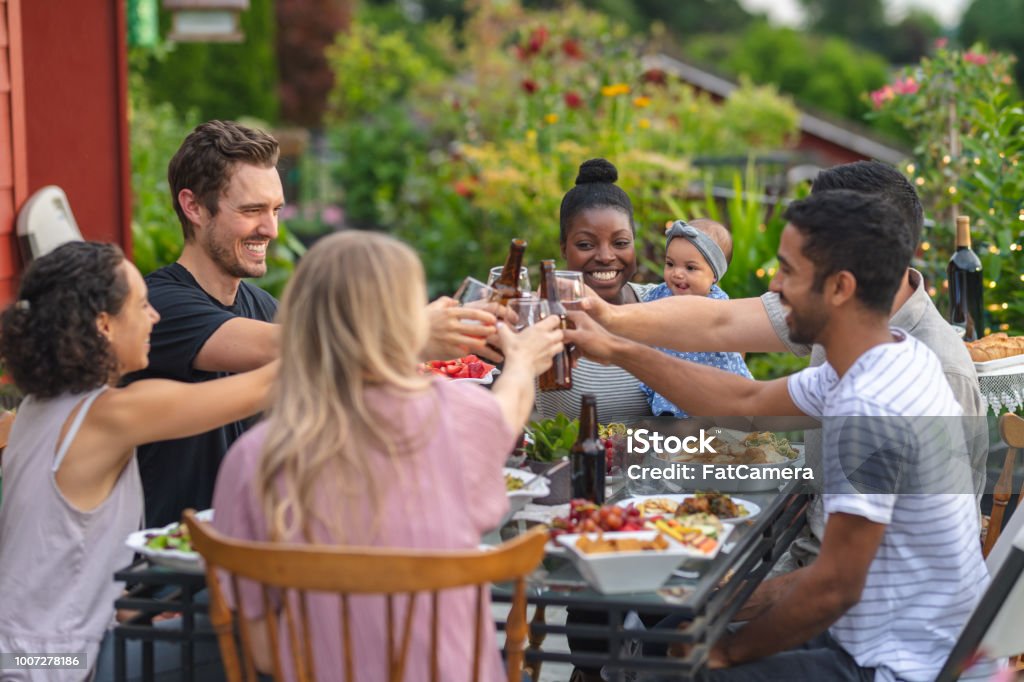 A group of young adult friends dining al fresco on a patio A multiethnic group of young friends enjoy good food and conversation together on a terrace outside on a summer evening. They're raising bottles and glasses up in a celebratory toast. Beer - Alcohol Stock Photo