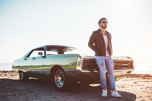 Handsome bearded man is standing near his green retro car on the beach. Vintage classic car.