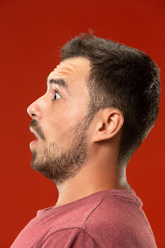 Wow. Attractive male half-length front portrait on red studio backgroud. Young emotional surprised bearded man standing with open mouth. Human emotions, facial expression concept. Trendy colors