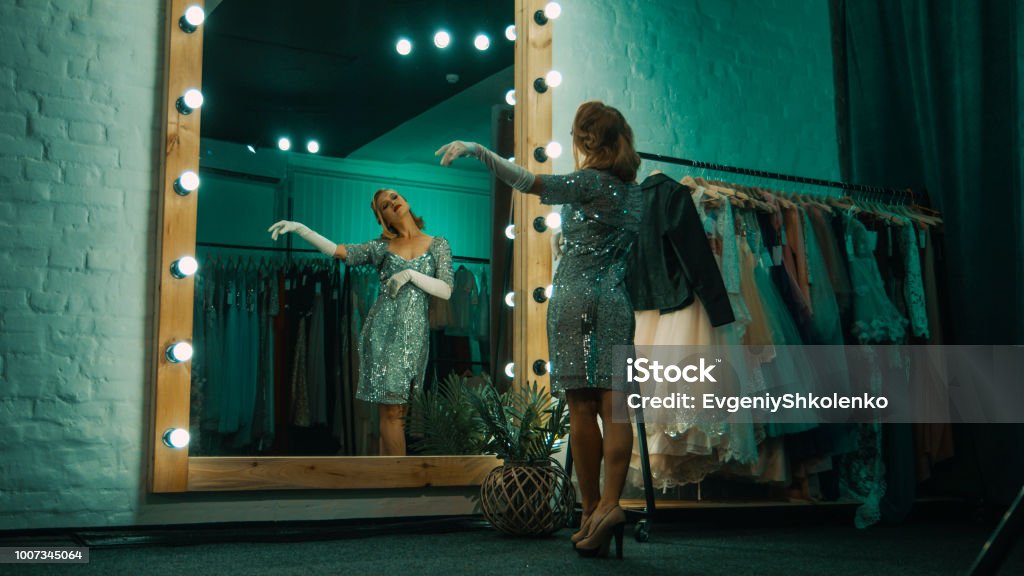 Elegant woman in dressing room before show From below back view of beautiful singer in short sparkling dress and gloves standing in front of mirror practicing show Theatrical Performance Stock Photo