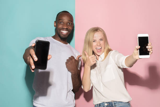 Portrait of a confident casual girl showing blank screen mobile phone and afro man Portrait of a confident casual girl showing blank screen mobile phone isolated over pink and blue background. Blond girl with freckles and afro man black men with blonde hair stock pictures, royalty-free photos & images