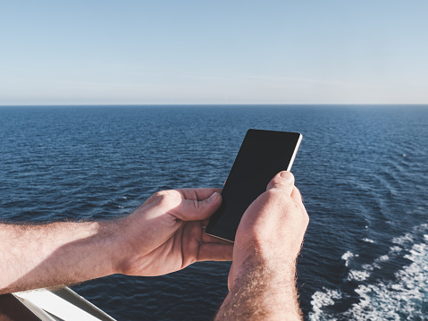 Men's hands holding a mobile phone on the deck of a cruise ship against the background of sea waves