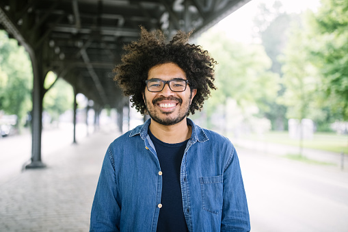 Portrait smiling african man wearing glasses. Man with curly hair standing under a city bridge and looking at camera.