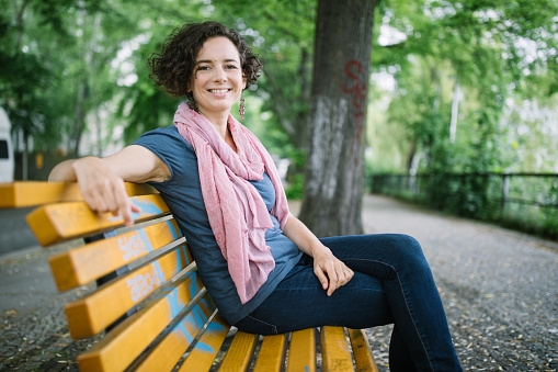 Beautiful woman sitting outside on park bench. Attractive female relaxing on bench outdoors.