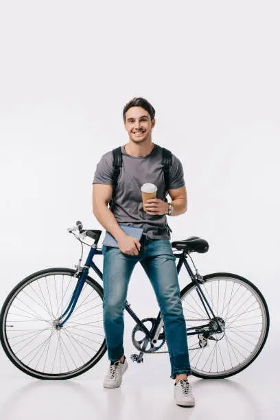 Photo of smiling student holding coffee to go and leaning on bicycle on white