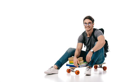 smiling handsome student sitting on skate with books on white