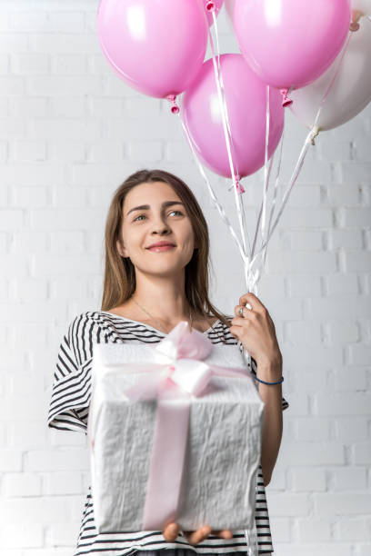 istock Smiling woman holding gift box and balloons on white brick wall background 1007425700