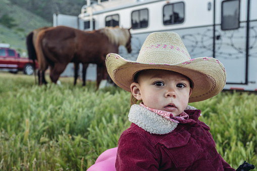 Portrait of a young Utah rancher, looking at camera. She is on the rocking horse, in the middle of  field with horses and trailer in the background..