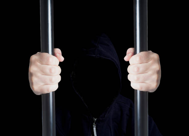 istock Anonymous criminal prisoner in hood holding prison bars 1007441962