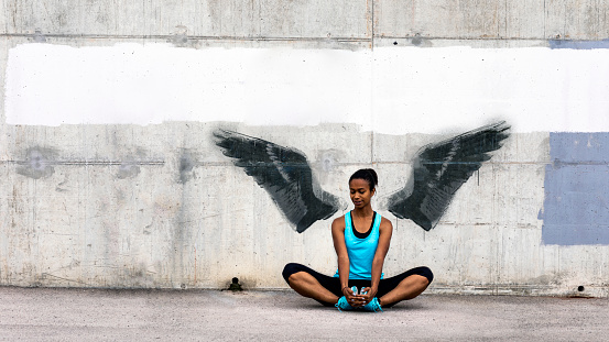 Young woman sitting on the floor, stretching in front of a concrete wall with wings graffiti. The graffiti is not real, the image of wings is my own.