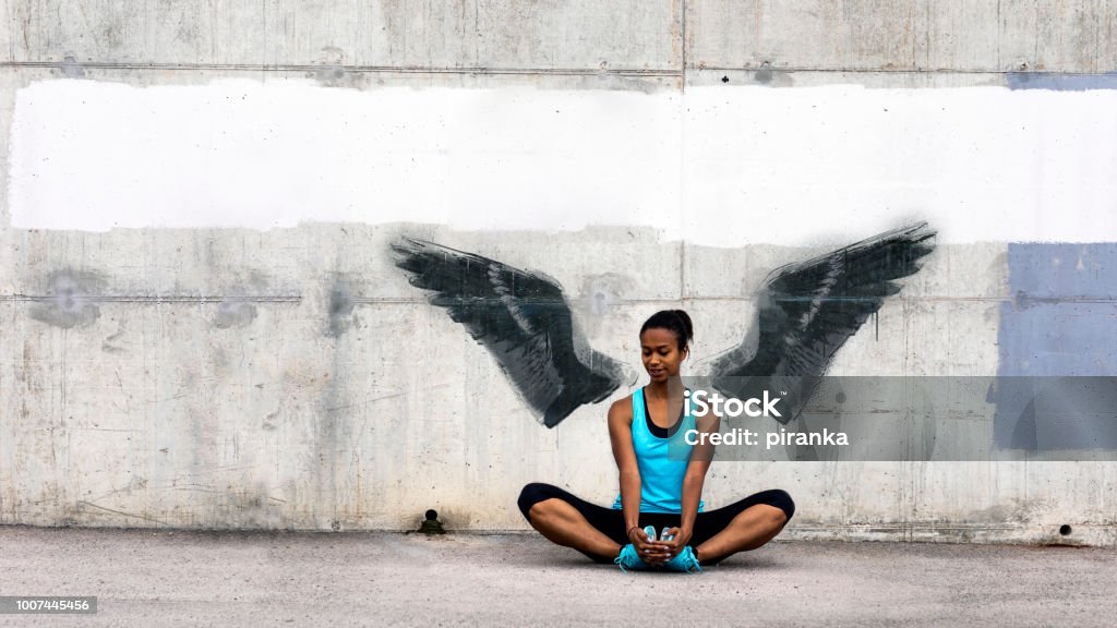 Female jogger Young woman sitting on the floor, stretching in front of a concrete wall with wings graffiti. The graffiti is not real, the image of wings is my own. Graffiti Stock Photo