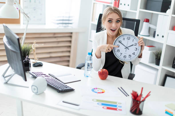 istock A young girl sits at a table in the office and points a finger at the clock face. Before the girl on the table there is an apple and there is a bottle of water. 1007448332