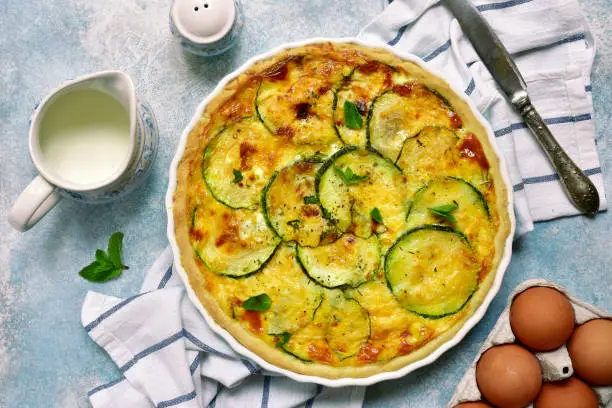 Delicious summer quiche with zucchini in a baking dish over light blue slate, stone or concrete background.Top view.