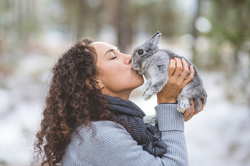 A young ethnic woman holds up her cute little bunny and kisses her. They're outside in the woods on a chilly winter afternoon.