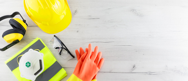 Banner image of construction safety gear including a hard hat, folded reflective jacket, face mask, goggles, and rubber gloves flat lay on white wooden surface with copy space