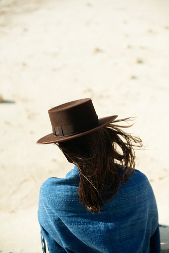 Brunette woman in brown hat and blue cloth standing in windy desert. Rear view.