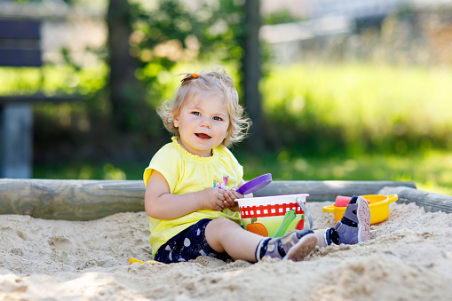 Cute toddler girl playing in sand on outdoor playground. Beautiful baby in red gum trousers having fun on sunny warm summer day. Child with sand toys