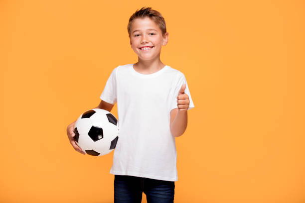 Happy little player hold soccer ball, smiling to the camera. A lot of copy space , yellow studio background.
