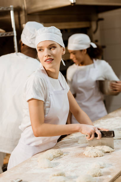 istock attractive female baker cutting dough with dough knife at baking manufacture while her colleagues working blurred on background 1007714114