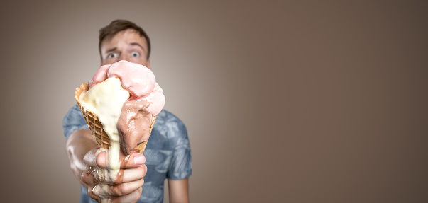 Young man in a Hawaiian shirt is holding a melting ice cream cone into the camera. With a funny / skeptical expression on his face. Isolated on a chocolate brown background. A lot of copy space.