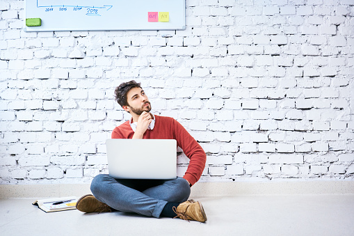 Male student sitting on floor with laptop and thinking. Portrait of startup entrepreneur