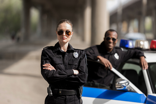 young serious policewoman standing with crossed arms while her partner standing near car and smiling blurred on background